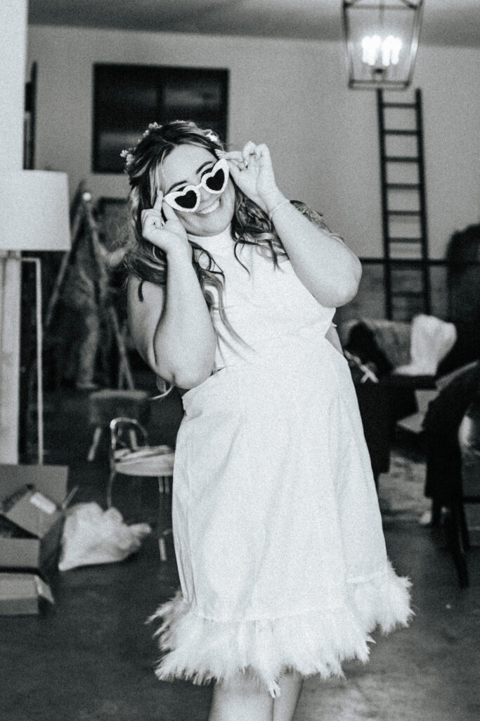 Bride smiles at photographer wearing heart shaped white sunglasses. Picture is an example of the blurry wedding photo trend.