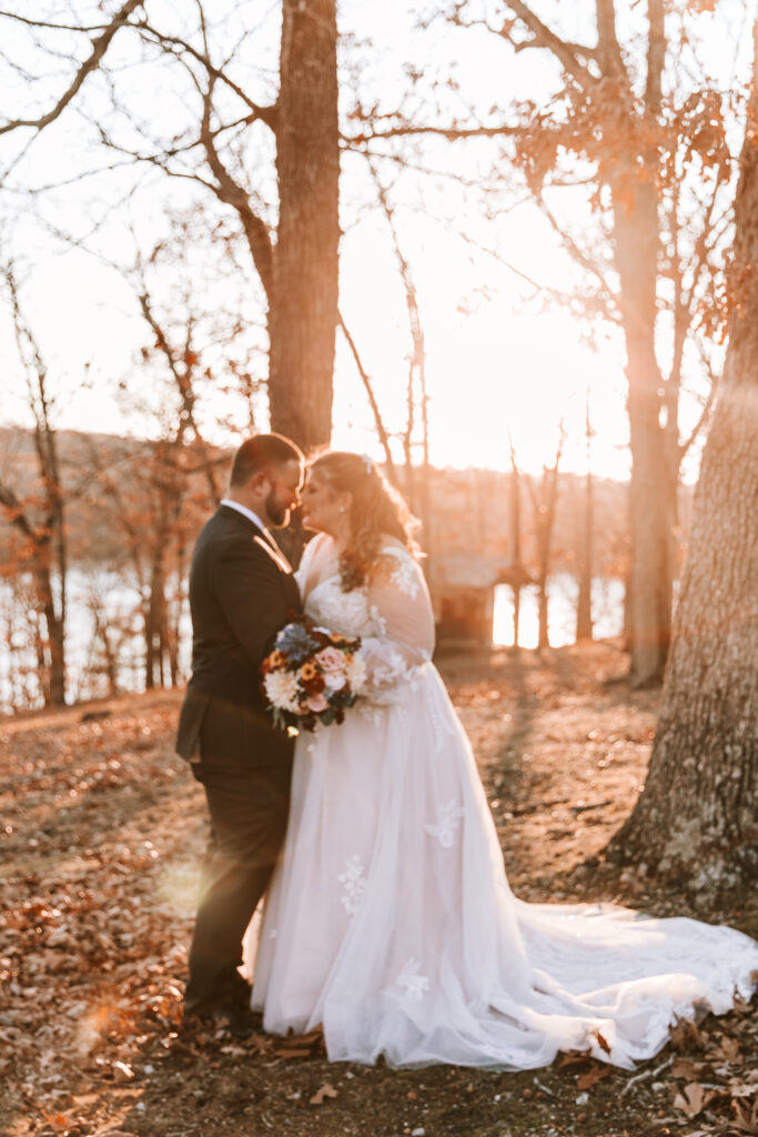 Groom and bride embracing in the woods of their wedding venue. There is a lake in the background and it is golden hour.