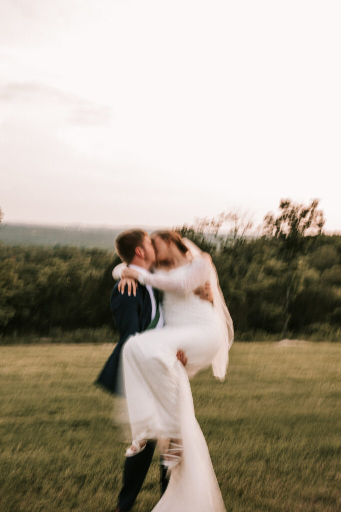 A groom carries his bride in the field of their wedding venue. The photo is in blurry wedding photo trend.