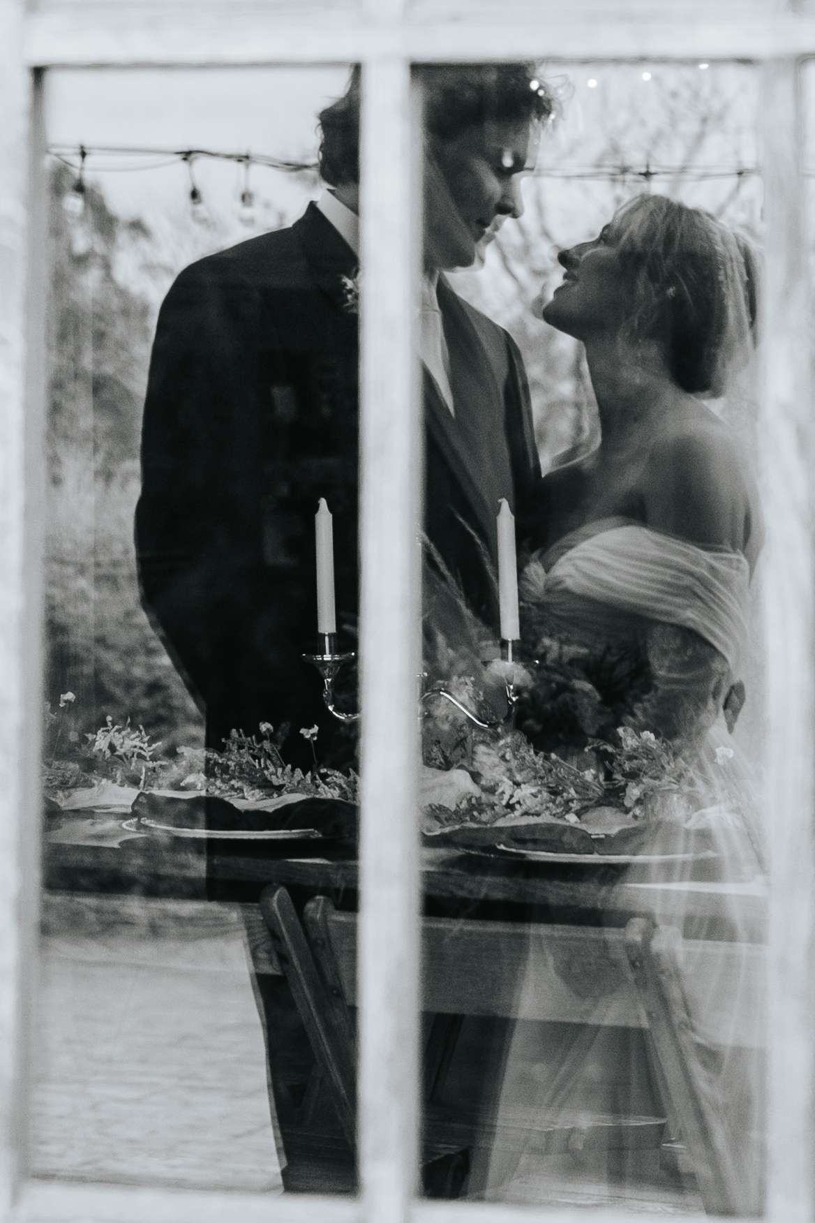 A groom looks at his bride reflecting off the window of the reception. The candle centerpieces and plates can be seen through the window. 