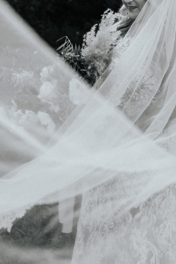 A bride's veil blows in the wind in front of her bridal bouquet. The photo is in black and white and is blurry which is a wedding photo trend.