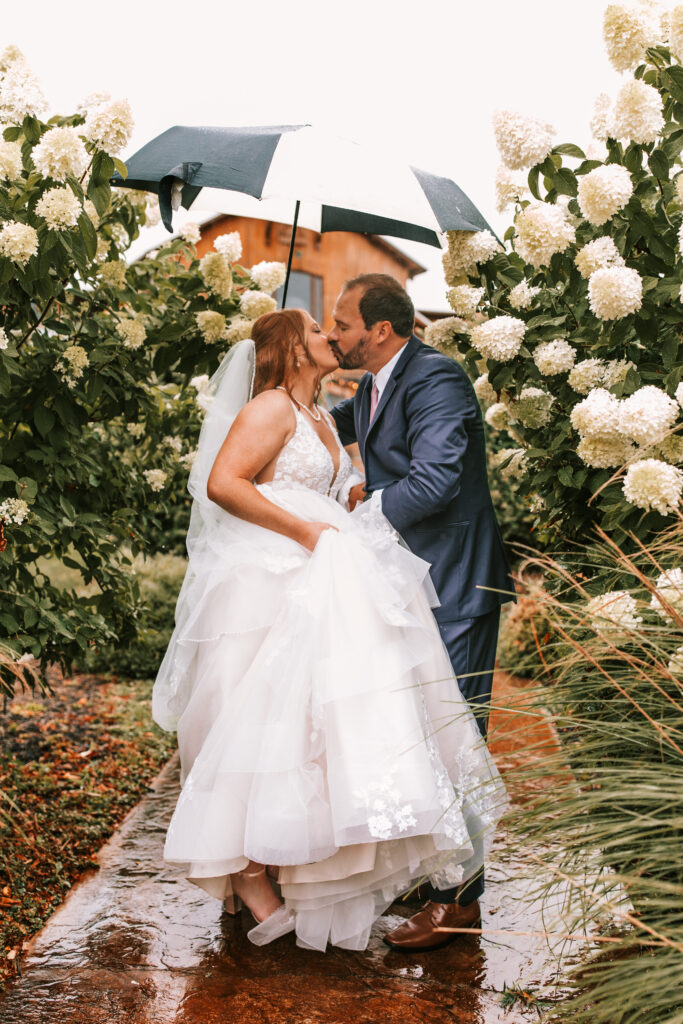 Bride and groom kissing under an umbrella between blooming flower bushes at Mighty Oak Lodge in lebanon Missouri. 

