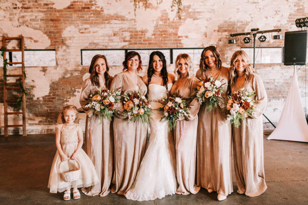 Spring season wedding Bridal party at Chrysler Commons Event Venue in Buffalo, Missouri standing in front of a distressed brick wall in champagne colored dresses.
