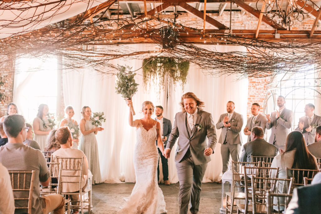 Newlyweds walking down the aisle after their ceremony at Chrysler Commons Event Venue in Buffalo, Missouri. Greenery and branches decorate wooden trusses with white fabric panels as the background.
