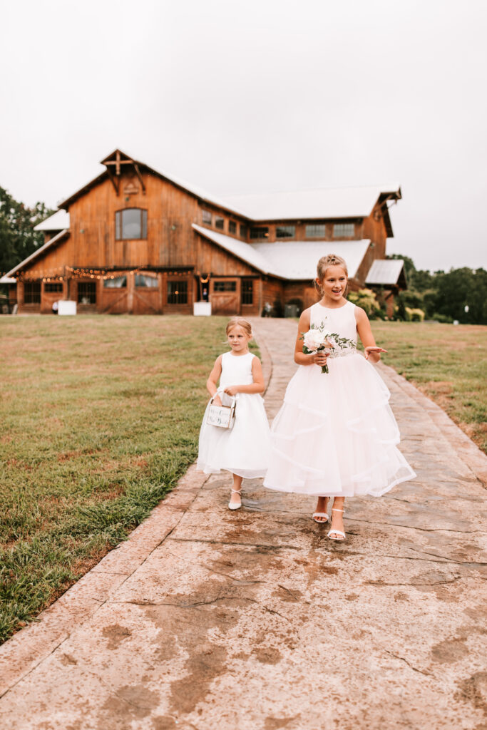 Two flower girls walking down the path from Mighty Oak Lodge to the oak tree for a ceremony. Girls are wearing white dresses and dropping flowers.