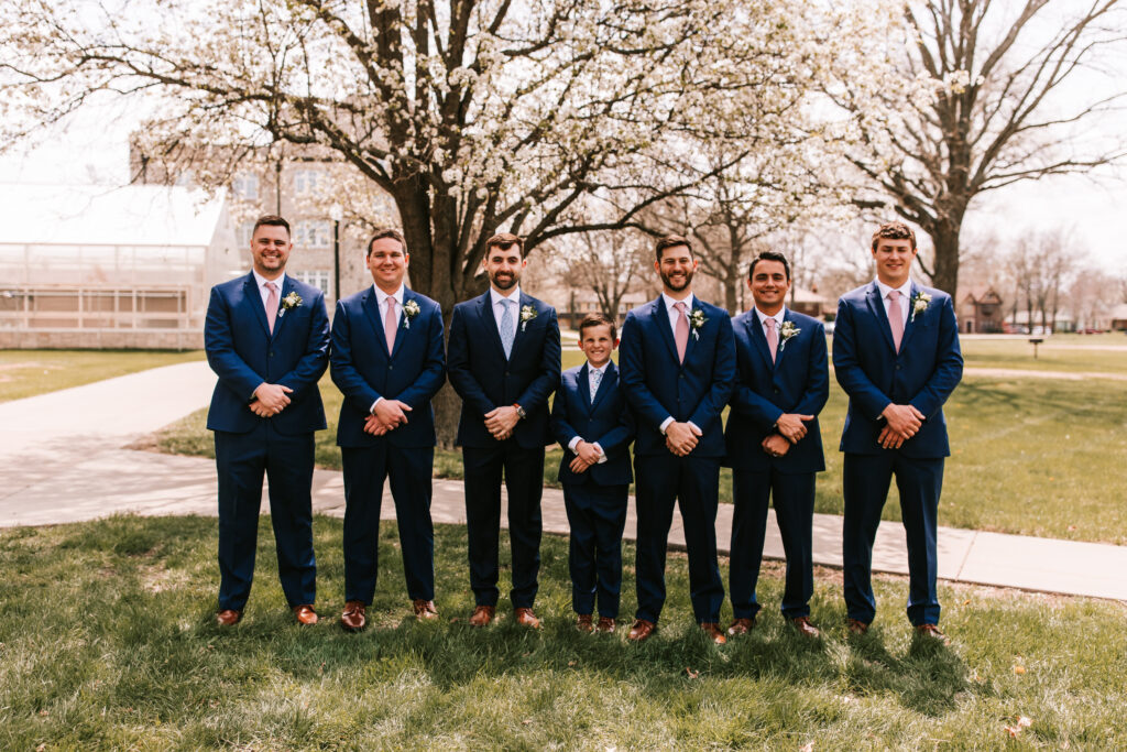 Dark blue suits on groomsmen standing outdoors at Fredrikson Center in Ottawa, Kansas at a spring season wedding photographed by Bailey Morris.