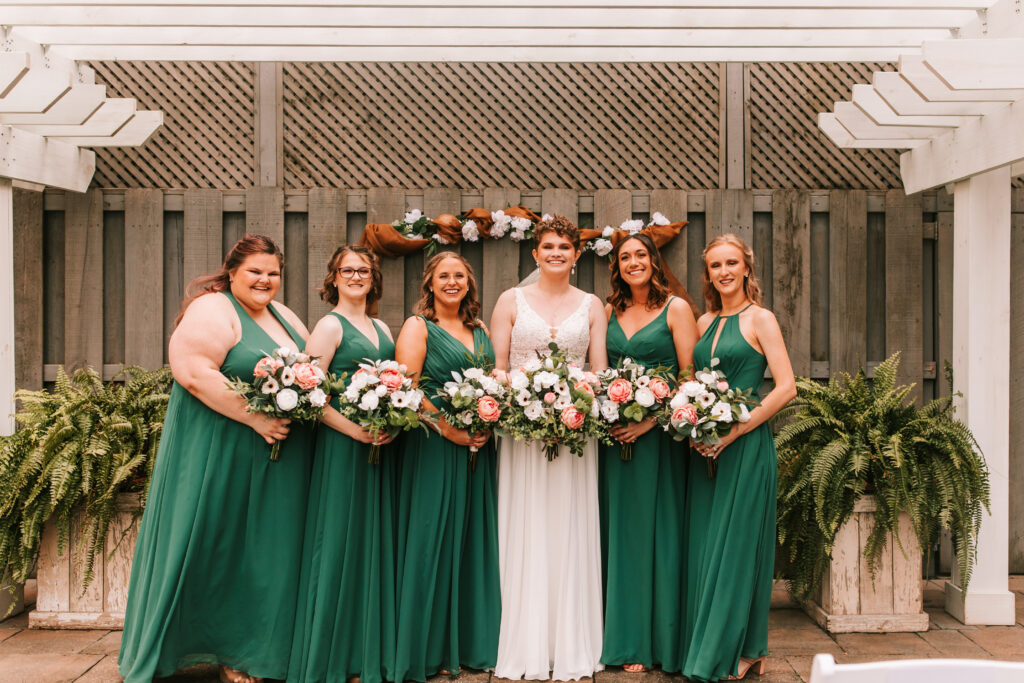 Bride with her bridesmaids wearing emerald green dresses holding peach and white flowers with lots of greenery in front of a textured, wooden fence.
