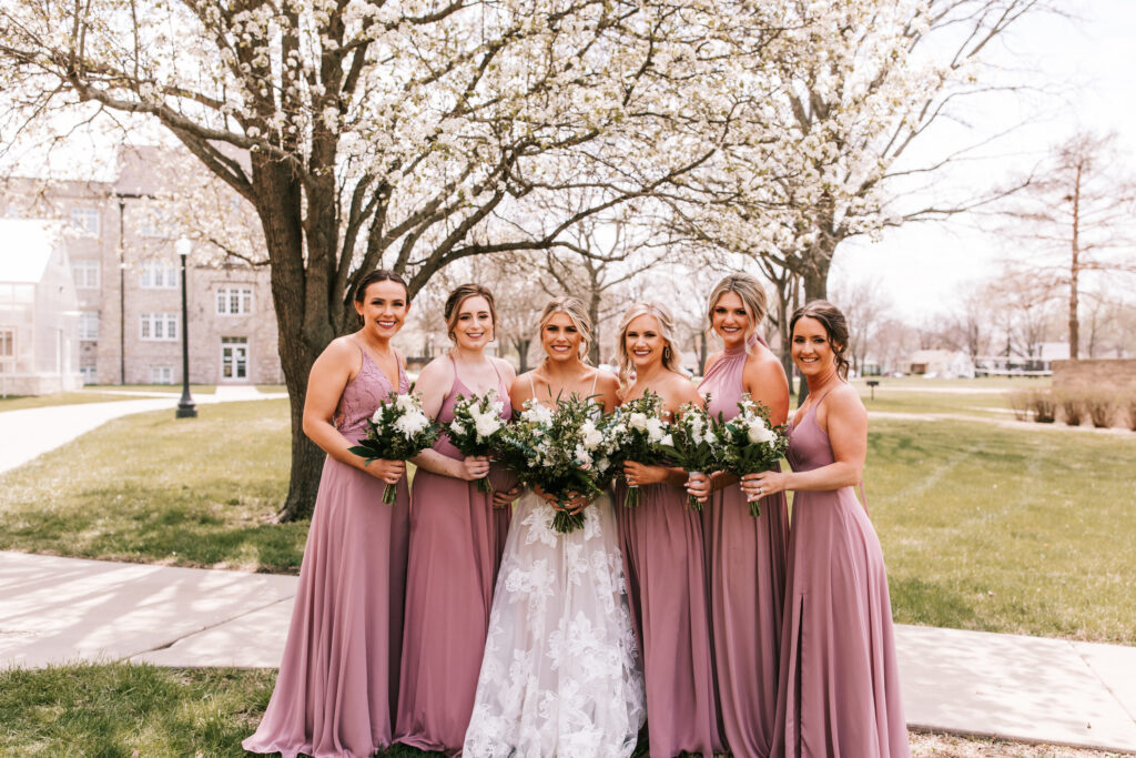Bride and bridesmaids at an outdoor ceremony spring season wedding wearing blush colored gowns photographed by Bailey Morris with trees blooming in the background.