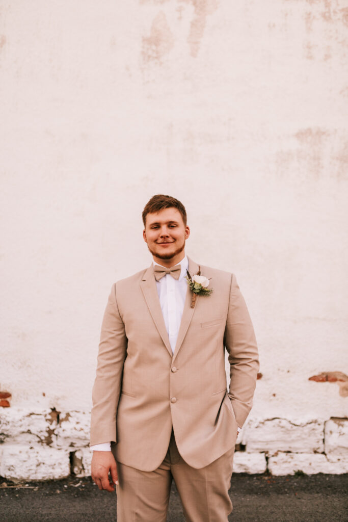 Groom in a spring season wedding khaki suit with a cream flower at a wedding venue in Buffalo, Missouri in front of distressed exterior wall photographed by Bailey Morris. 