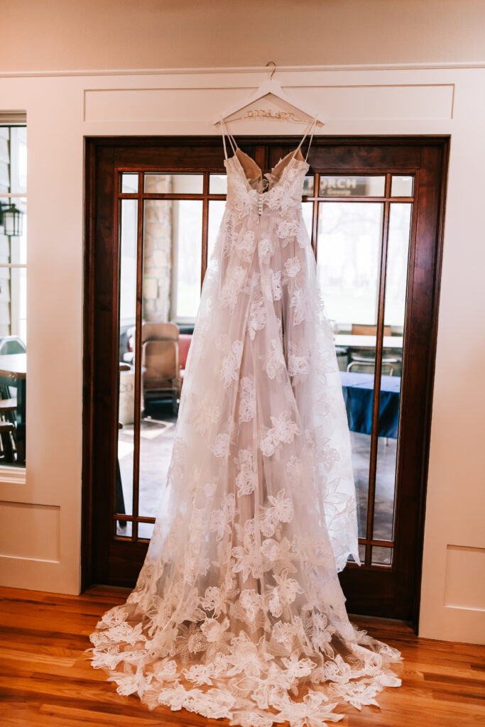 Floral lace wedding gown hanging from hanger in front of a glass door with wooden frame spreading out over a hardwood floor in Kansas City, Missouri.
