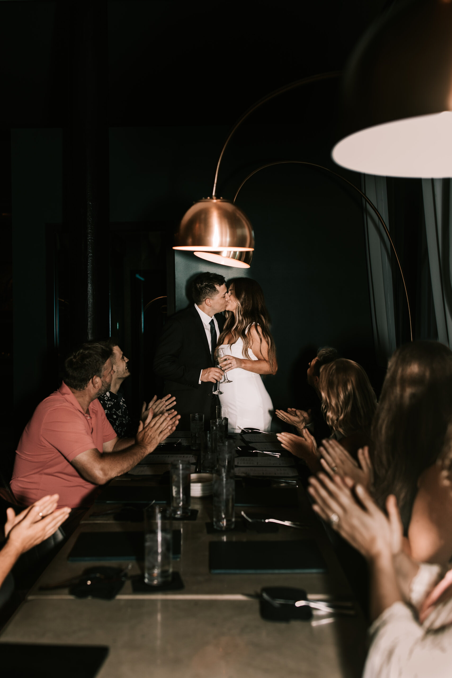 Bride and groom kiss after a toast at their Hotel Vandivort wedding while their guests look on. Gold pendant lights hang in the dark bar. 