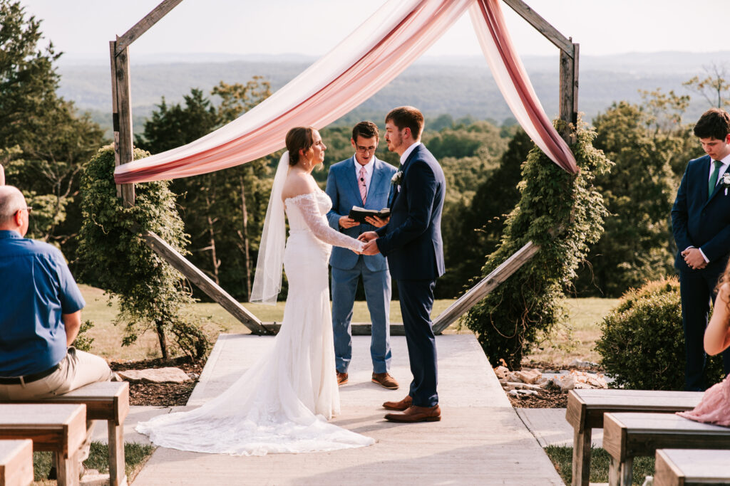Couple saying their vows in front of a wooden octagon with a blush colored sash outdoors at The Atrium wedding venue in Branson, Missouri.