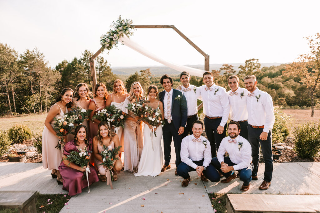 Wedding party in front of wooden octagon arbor with floral decor and white sash. Branson wedding venue: The Atrium. Branson wedding photographer: Bailey Morris.