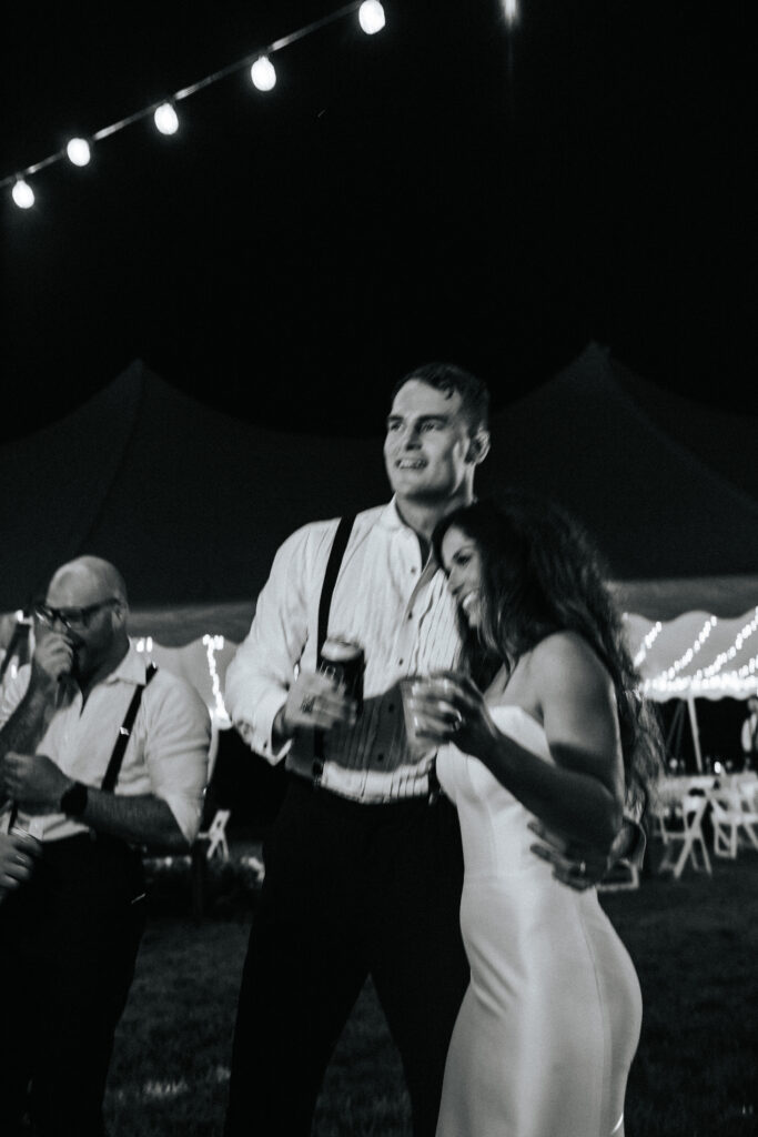 Groom and bride holding a drink, hugging, and admiring the guests having fun at their wedding reception. Black and white wedding photos by Bailey Morris.