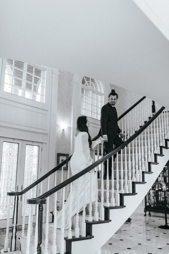 A groom leads his bride up a beautiful staircase at their wedding in Lebanon, Missouri. Black and white wedding photos by Bailey Morris.