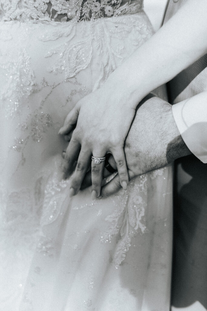 Bride has her hand placed over her grooms hand and her wedding ring is showing. Both hands are on top of her lace wedding gown. Black and white wedding photos by Bailey Morris.