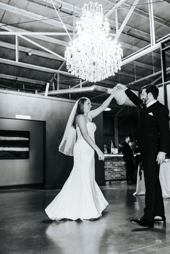 A groom is twirling his bride on the dance floor under a chandelier at a wedding venue in Missouri.Black and white wedding photos by Bailey Morris.