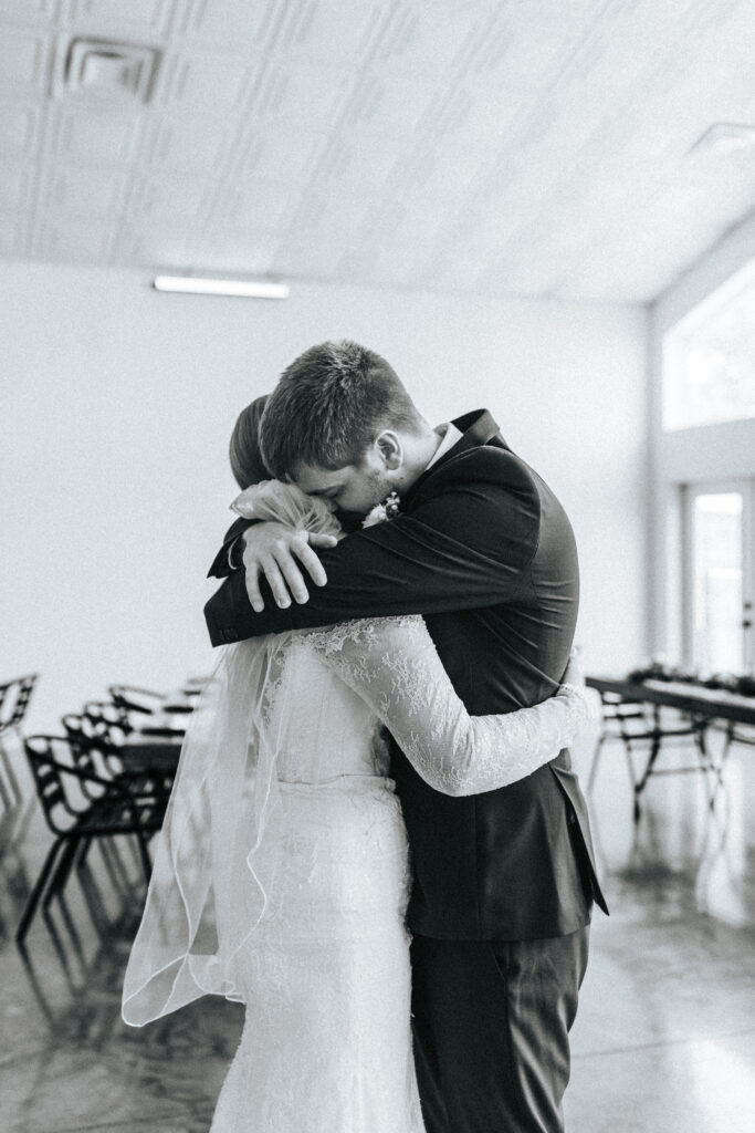 A groom embraces his wife with his face down into her neck and shoulder at their Missouri wedding Venue. Black and white wedding photos by Bailey Morris.