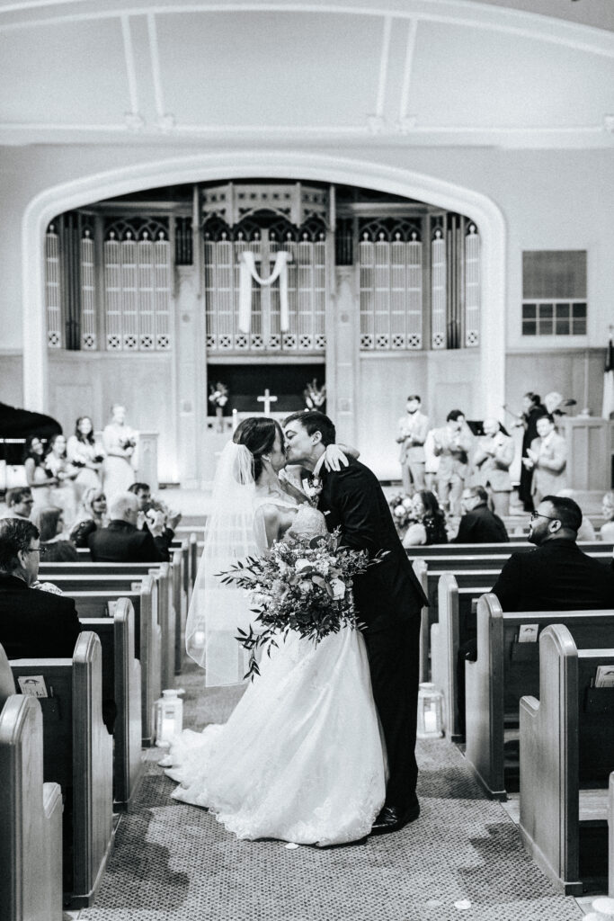 A groom kisses his bride in the aisle as they exit the church. Guests cheer for them from the pews at a Missouri wedding. Black and white wedding photos by Bailey Morris.