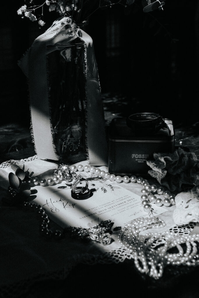 The ring, pearls, and wedding invitation are lying on a table. There are flowers in a vase and their is a shadow cast across the photo. Black and white wedding photos by Bailey Morris.