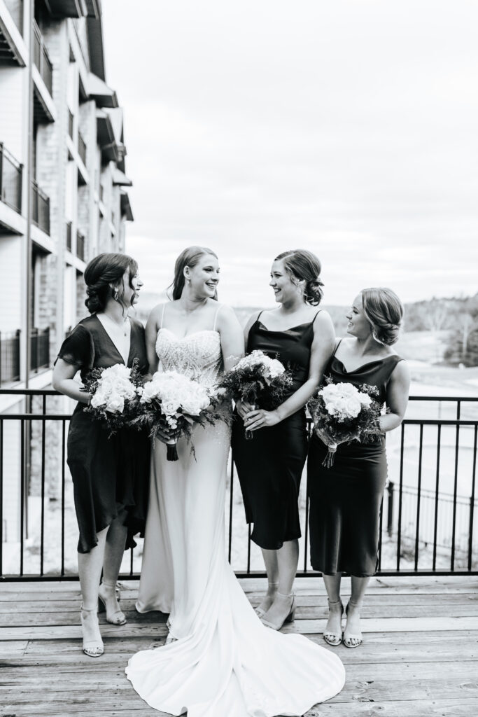 A bride and 3 of her bridesmaids stand on a balcony at their Missouri wedding venue. The women are all holding their bridal bouquets. Black and white wedding photos by Bailey Morris.