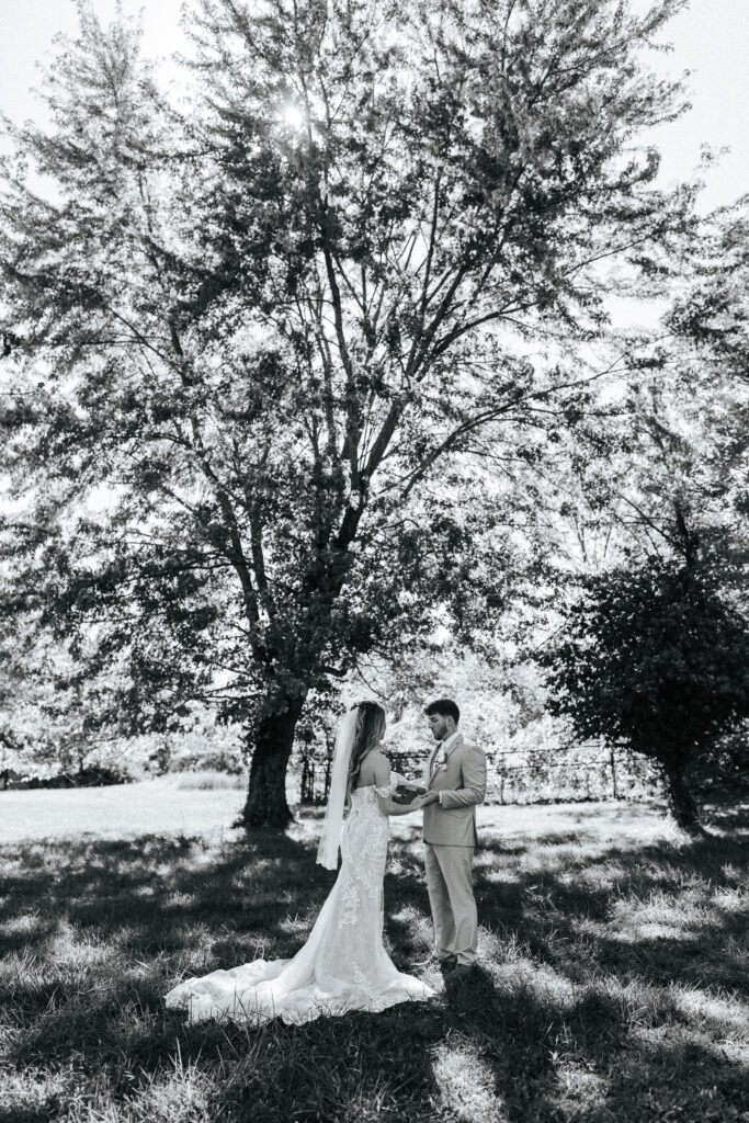 A bride and groom hold hands beneath a large tree at their Missouri wedding venue. Black and white wedding photos by Bailey Morris.