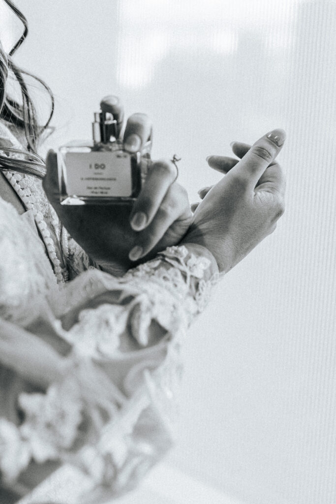 A bride in a long sleeved wedding gown sprays a bottle of perfume on her wrist. Black and white wedding photos by Bailey Morris.