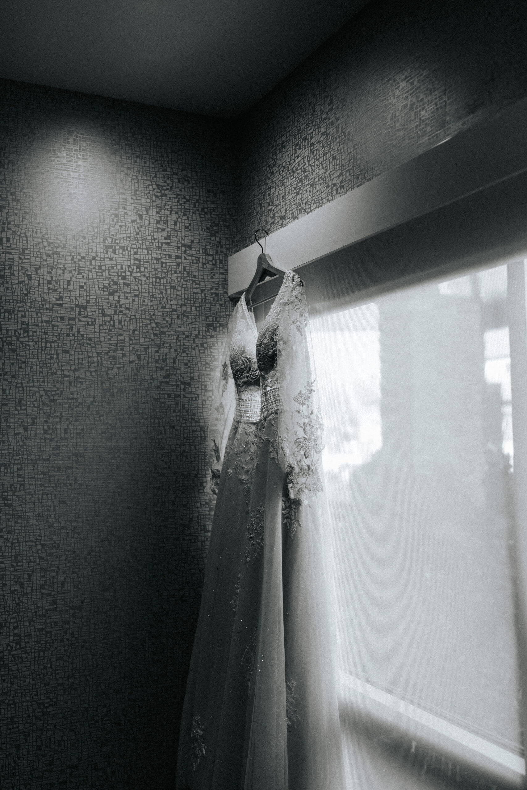 Black and white photo of wedding dress hanging from a hanger in front of a window in a bridal suite of a Missouri wedding venue. Photo by Bailey Morris wedding photographer.