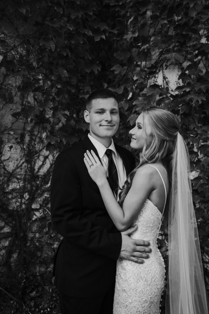 Black and white photo of bride looking at groom while groom looks at camera. Couple are standing in front of vine covered wall at Haseltine Estate in Springfield, Missouri.