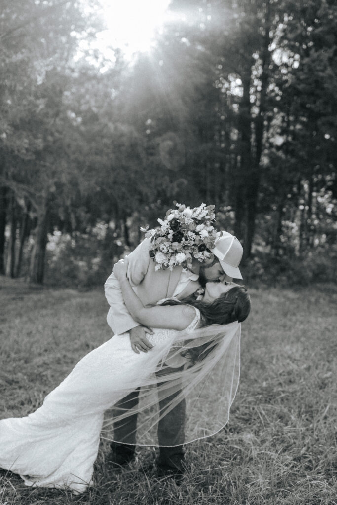 A groom dips his bride for a kiss as she rests her bouquet on his back in a field at a Missouri wedding venue. Black and white wedding photos by Bailey Morris.