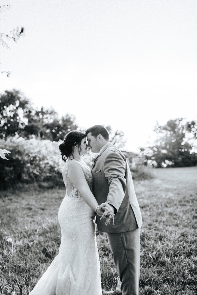 A bride and groom interlock hands with their foreheads touching in a field at a Missouri wedding venue. Black and white wedding photos by Bailey Morris.