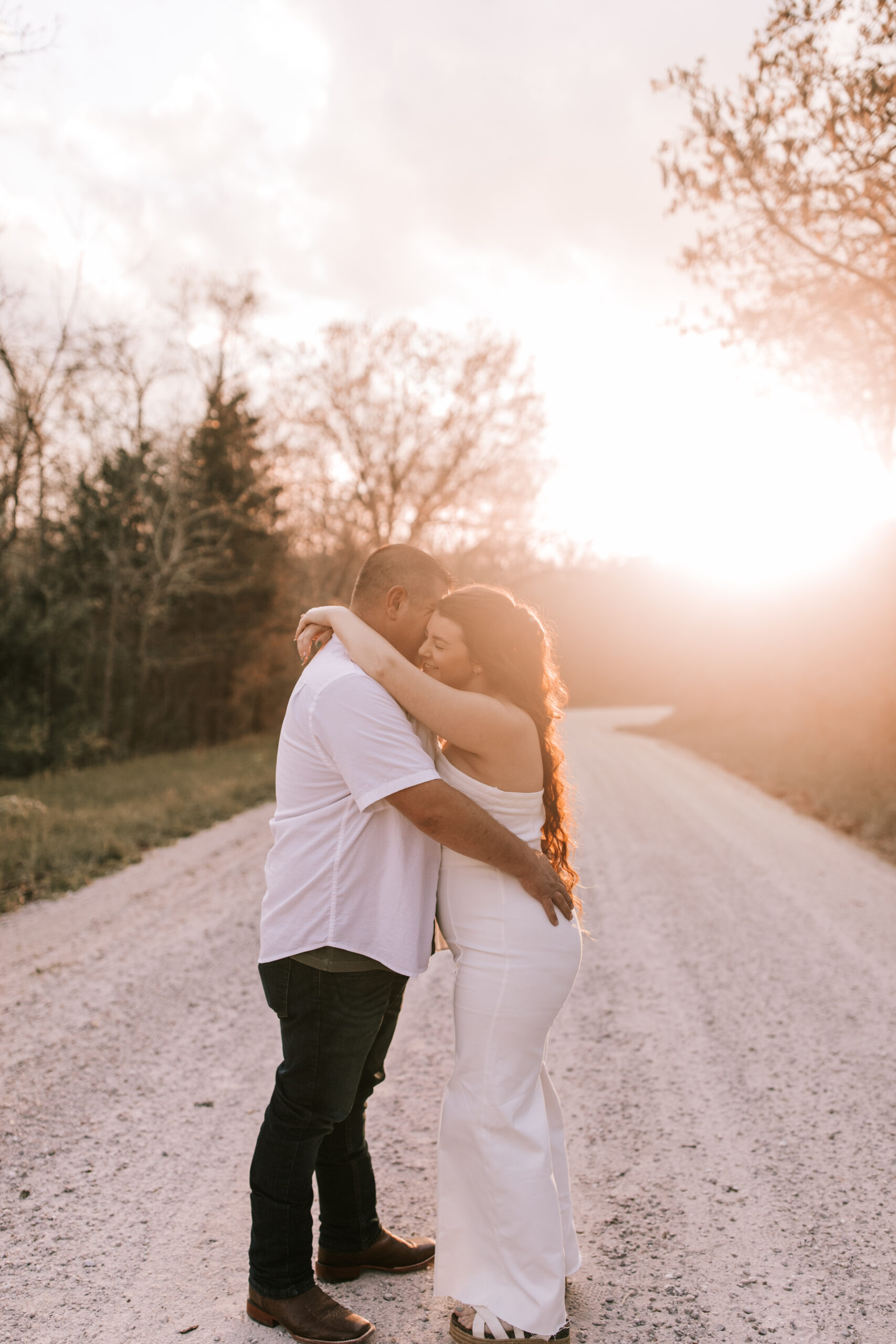Missouri engagement photo. Couple is standing on a gravel road with the sun setting in the background. She is wearing a white dress and he is wearing a white shirt and jeans. Photo by Bailey Morris wedding photography.