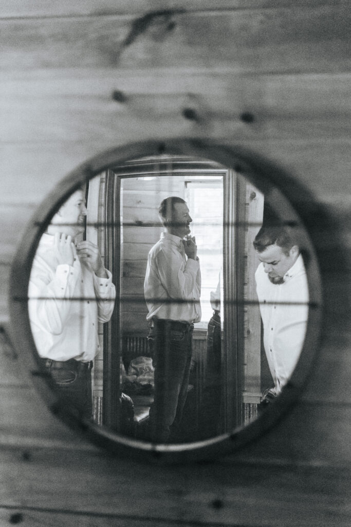 A circular mirror on a wood paneled wall reflects a groom tying his tie in preparation for his Missouri wedding. Black and white wedding photos by Bailey Morris.