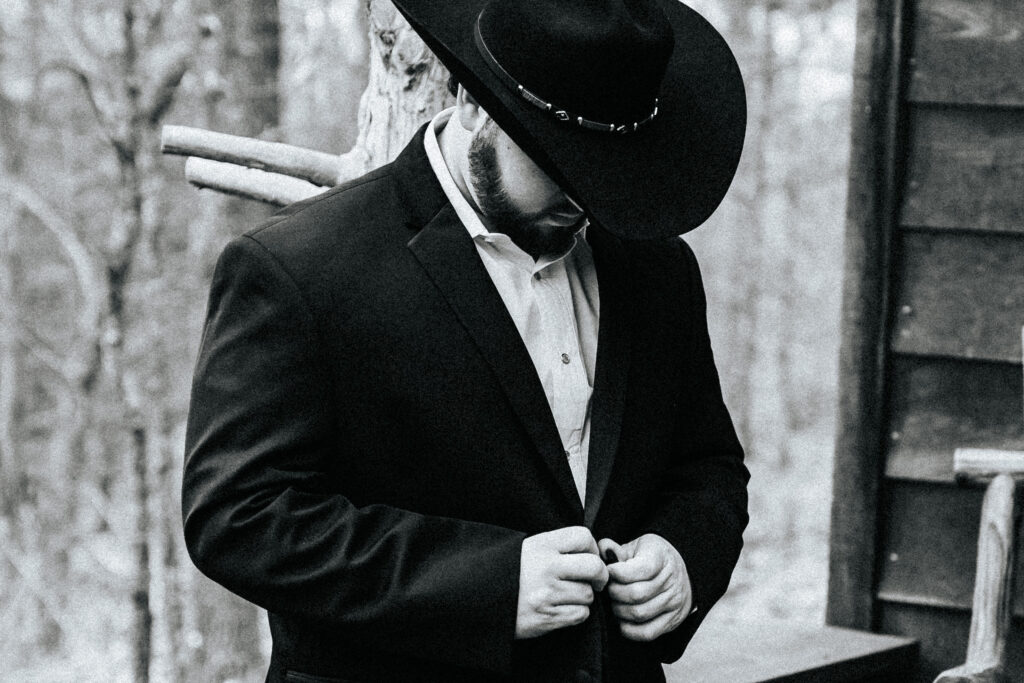 A groom in a black cowboy hat looks down as he buttons his blazer before his Missouri wedding. Black and white wedding photos by Bailey Morris.