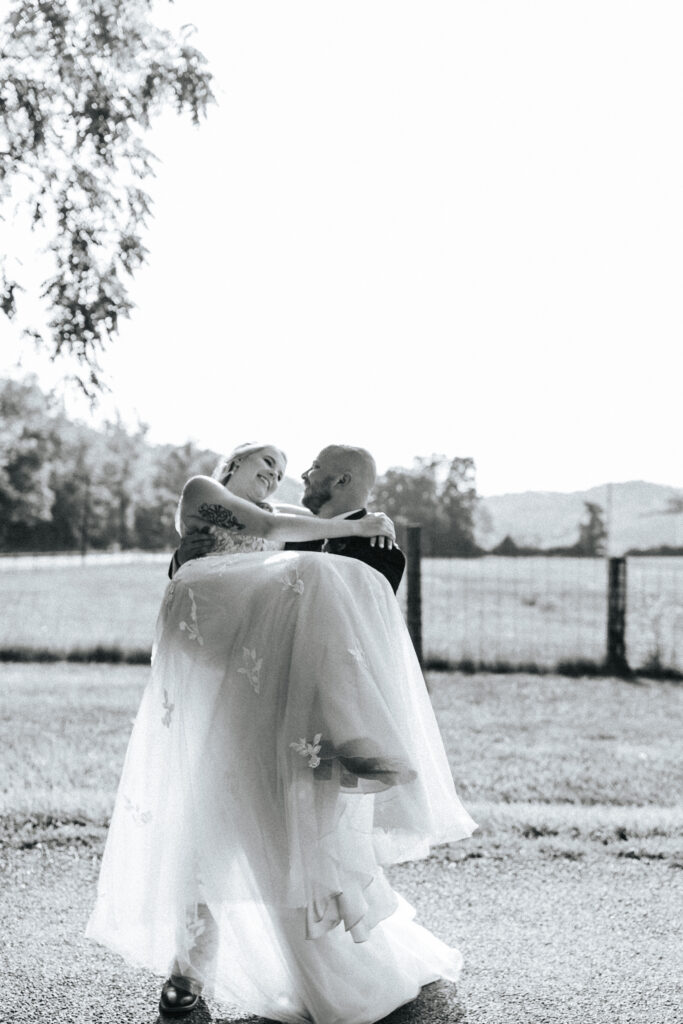 A groom carries his bride through a field at their Missouri wedding venue. Black and white wedding photos by Bailey Morris.