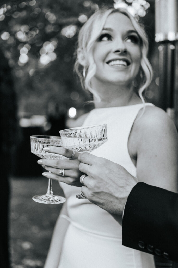 A bride smiles as she clinks crystal glasses with her groom for a toast at their Lebanon, Missouri wedding. Black and white wedding photos by Bailey Morris.