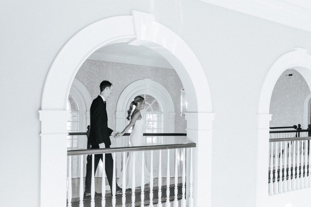 A bride leads her groom down the hallway. The hallway features beautiful large arch windows and cutouts as well as a wooden staircase and railings. Black and white wedding photos by Bailey Morris.