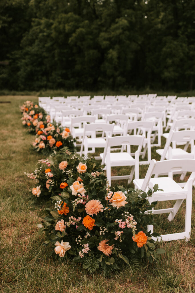 Wedding florals by Carley Jean Floral in Jefferson City Missouri. Arrangements line the aisle between two sets of white wooden chairs set up in a field for a farm wedding. 