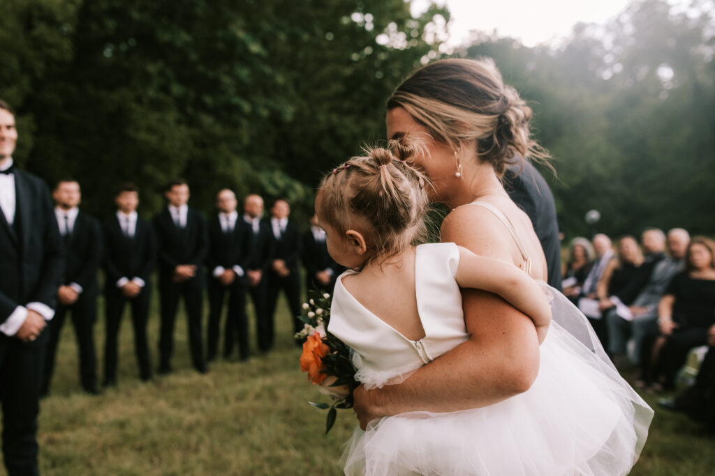 Bridesmaid carries flower girl down the aisle in front of the groomsmen on a farm wedding in Jefferson City Missouri. Photo by Bailey Morris Wedding Photographer.