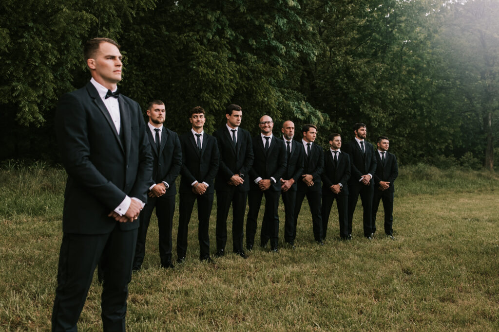 Groom and his groomsmen waiting for the wedding to start in a field in front of dense woods. Bailey Morris Jefferson City Wedding Photographers.
