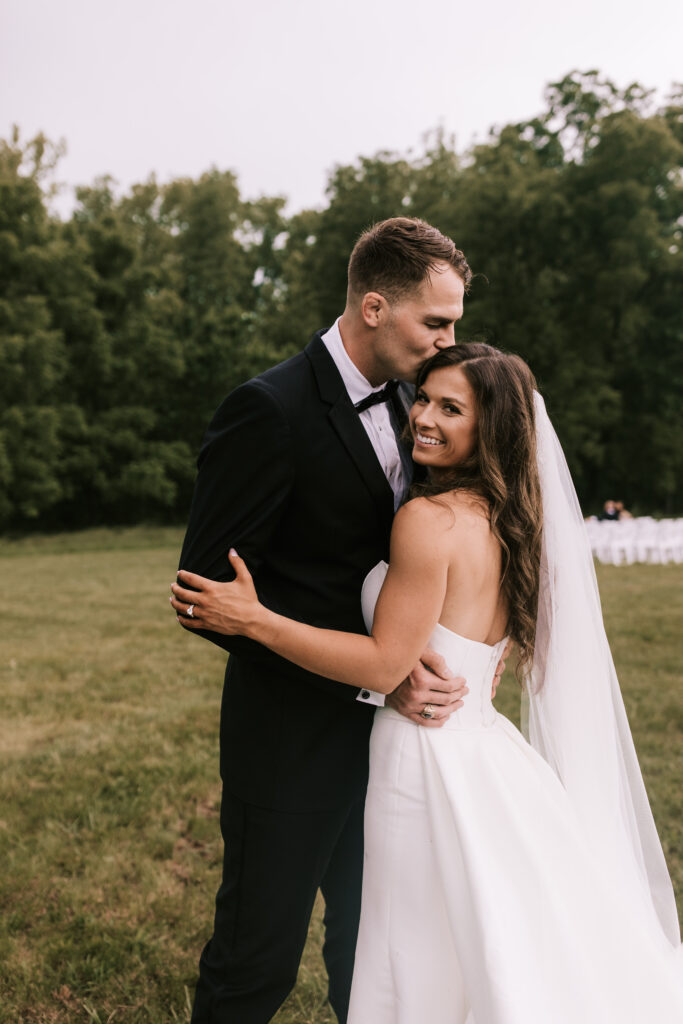 Groom kissing bride on the head in front of their ceremony site on a field on private property in Jefferson City Missouri.