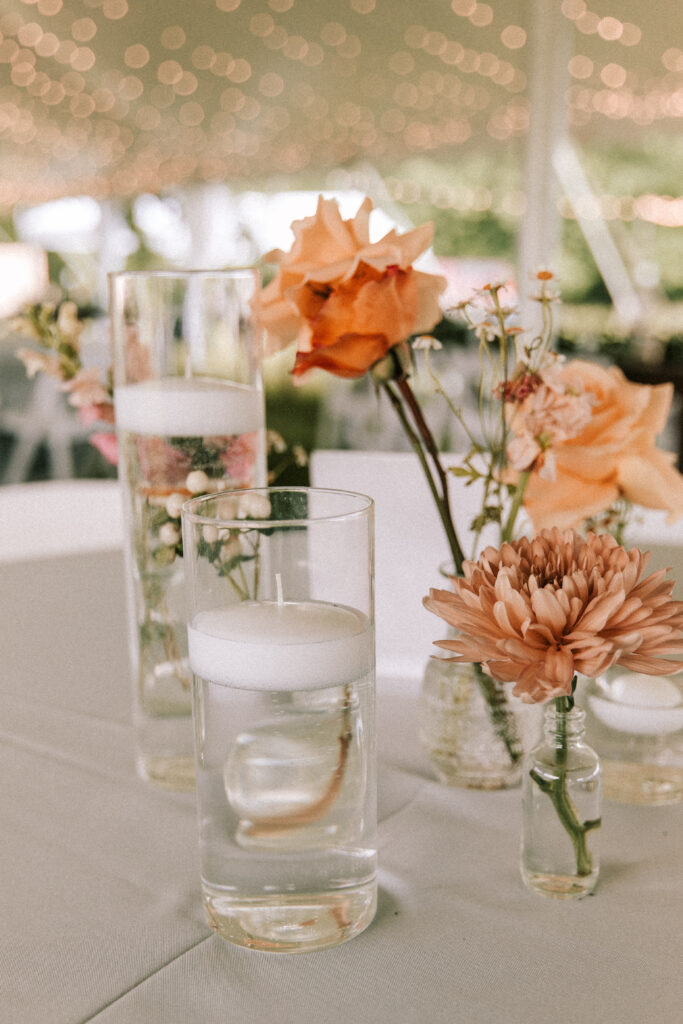 Small vases with coral and peach flowers on a table at a wedding reception in Jefferson city Missouri. There are cylindar glasses with floating candles as an additional accessory under the tent with string lights. 
