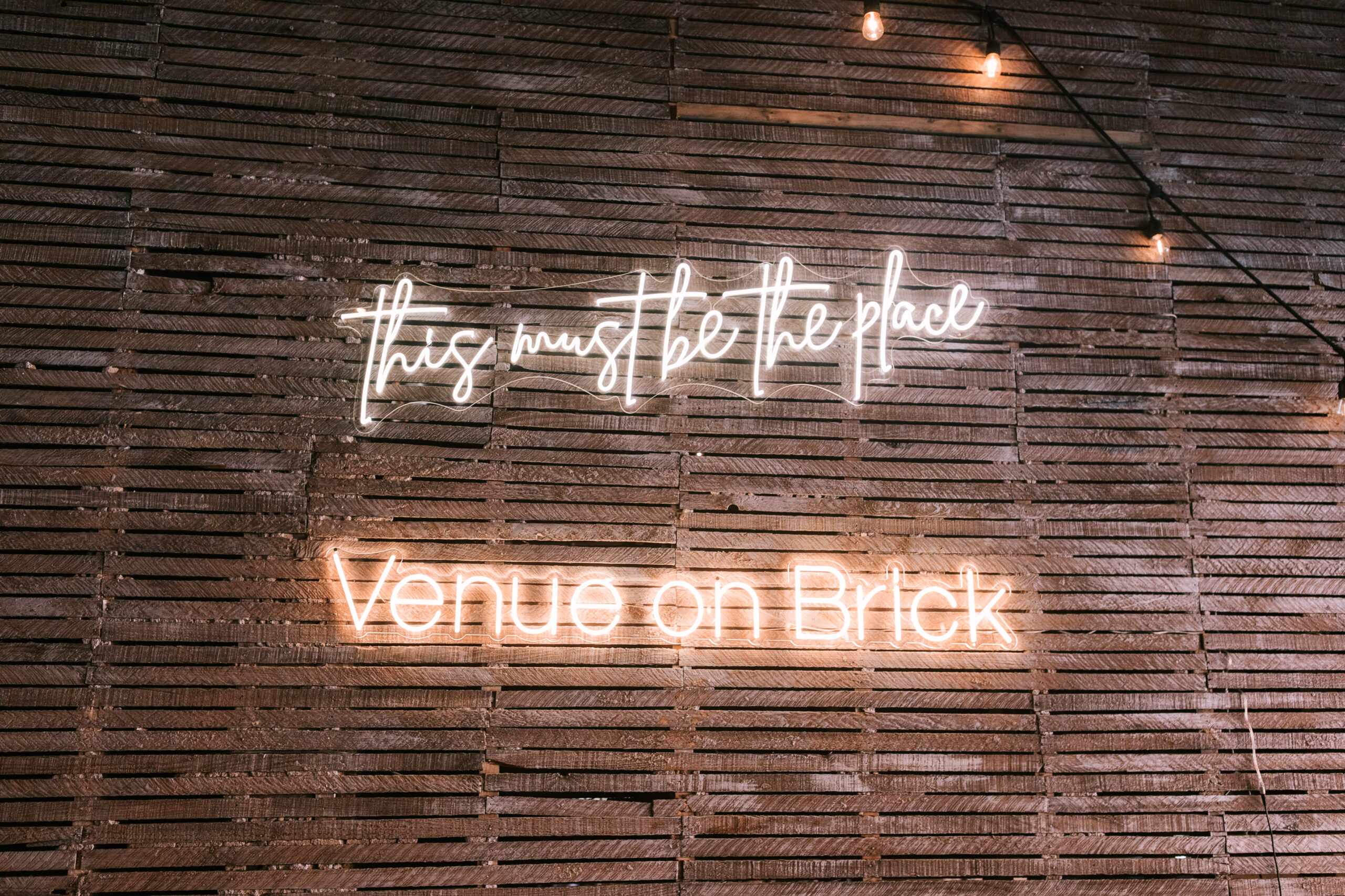 Neon letters spell out "this must be the place Venue on Brick" against an old wooden, distressed wall.