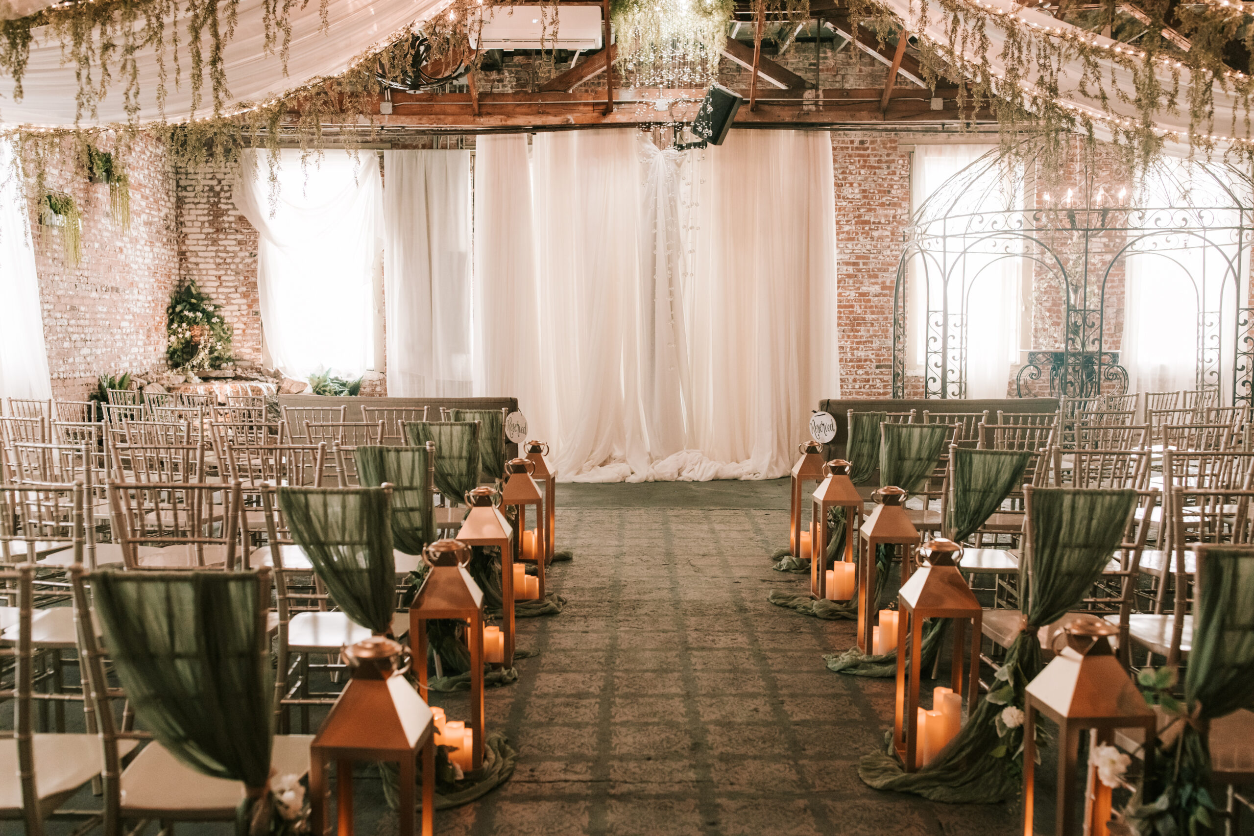 Ceremony space at Venue on Brick includes draped white material, greenery, twinkle lights, and a chandelier against a wall of exposed brick. Copper lanterns line the aisles.